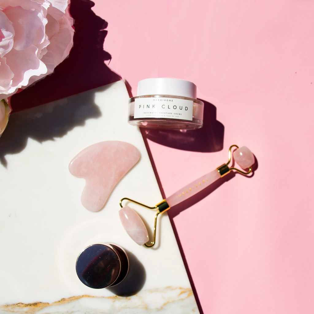 A photo of pink quartz skin roller with dual ends, a gua sha stone, a jar of moisturizer, eye cream, and flowers. The image is of products used in the blog post "The Skinny on Skin Tools"